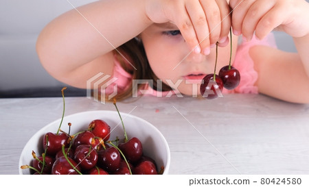 little girl sitting at a light wooden table with a plate of cherries and holding red berries 80424580