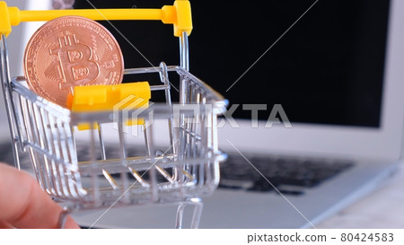 Small shopping cart with gold Bitcoin on it on the background of a laptop. Suitable for concepts as 80424583