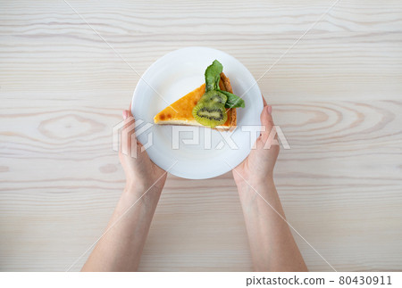 Delicious dessert. Woman holding plate with slice of cheesecake decorated with kiwi slices, wooden table, top view 80430911
