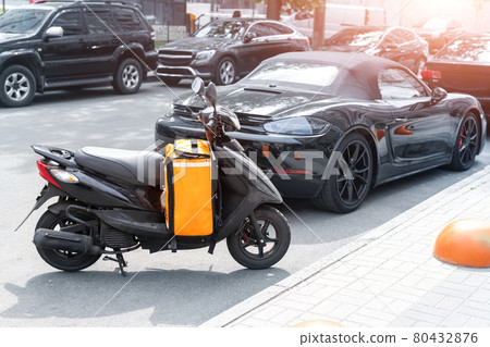 Fast express takeaway online gig order food delivery scooter with red orange bag box parked at traffic city center downtown street on sunny day. Eat quick moto courier shipment service transport 80432876