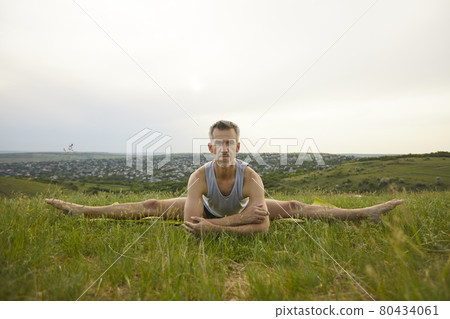 Serious male yoga instructor practicing stretching leg exercise in green summer meadow 80434061