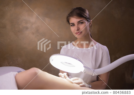 Female legs on purple sheet during epilation by a professional beautician in gloves. Spa, beauty industry, treatment at clinic, electrolysis. Female legs on purple sheet during epilation by a professional beautician in gloves. Spa, beauty industry, treatment at clinic, electrolysis. 80436883