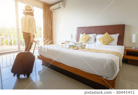 Portrait of tourist woman looking to beautiful view outside the window with her luggage in hotel bedroom after check-in. Conceptual of travel and vacations. Portrait of tourist woman looking to beautiful view outside the window with her luggage in hotel bedroom after check-in. Conceptual of travel and vacations. 80445451