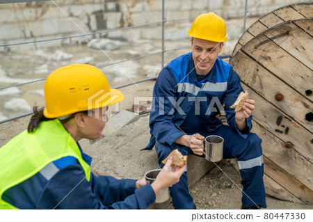 Two builders in hardhats and workwear having sandwiches with tea 80447330