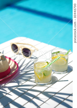 lemonade in glasses with lemon slice, straw hat and sunglasses on white table, edge of the pool with turquoise blue water background , light and shadows, vocation and summertime concept. 80448700