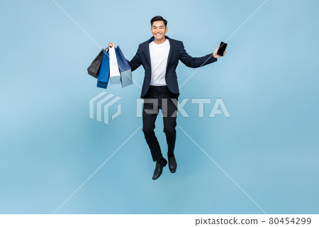 Excited young handsome Asian man holding mobile phone and shopping bags jumping Excited young handsome Asian man holding mobile phone and shopping bags jumping 80454299