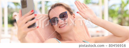 Portrait of smiling woman lying on sun lounger on beach and waving her hand into smartphone Portrait of smiling woman lying on sun lounger on beach and waving her hand into smartphone 80456916