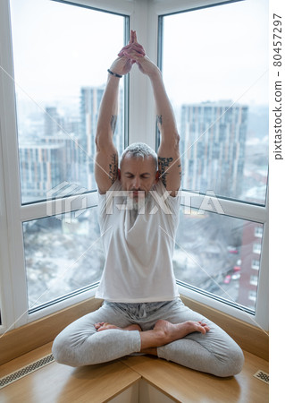 Gray-haired man in a white tshirt doing yoga and looking concentrated Gray-haired man in a white tshirt doing yoga and looking concentrated 80457297