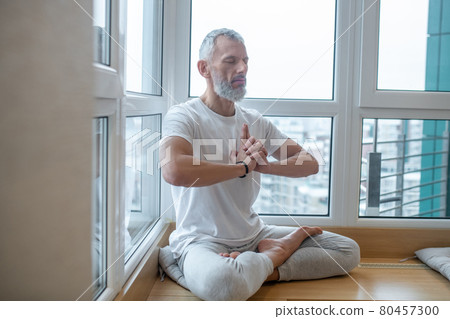 Gray-haired man in a white tshirt doing yoga and looking concentrated Gray-haired man in a white tshirt doing yoga and looking concentrated 80457300