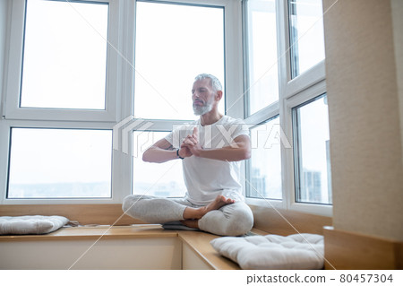 Gray-haired man in a white tshirt doing yoga and looking concentrated Gray-haired man in a white tshirt doing yoga and looking concentrated 80457304