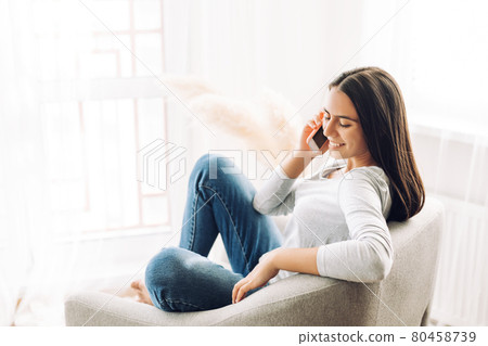 A young girl is talking on a mobile phone and sitting in a chair, climbing into it with her feet A young girl is talking on a mobile phone and sitting in a chair, climbing into it with her feet 80458739