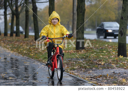 Gloomy boy in a yellow raincoat rides a bicycle along a wet alley in the rain. Autumn day 80458922
