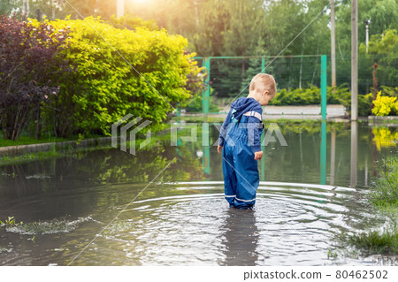 Little cute playful caucasian blond toddler boy enjoy have fun playing jumping in dirty puddle wearing blue waterproof pants and rubber rainboot at home yard street outdoor. Happy childhood concept 80462502