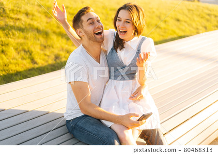 Medium shot portrait of laughing young couple hugging sitting on bench in city park in summer sunny day. 80468313