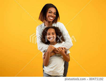 Mother's love. Happy loving african american mom cuddling her daughter from the back, standing over yellow background Mother's love. Happy loving african american mom cuddling her daughter from the back, standing over yellow background 80471192