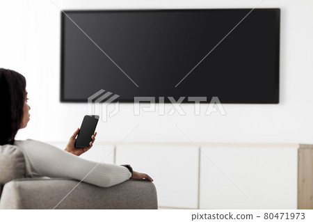 Modern gadgets. Black lady with smartphone connected to TV, watching video at home, plasma television with empty screen 80471973