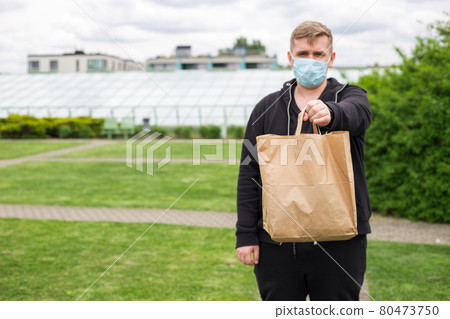 Close up of man hand in mask holding paper bag for takeaway food on nature green background. Delivery in any weather around the clock to the client. Close up of man hand in mask holding paper bag for takeaway food on nature green background. Delivery in any weather around the clock to the client. 80473750