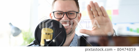 Smiling man sits in front of a microphone and waves at camera with his hand Smiling man sits in front of a microphone and waves at camera with his hand 80474859