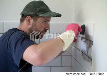 Worker applies grey grout at white tiles with rubber trowel. 80477084