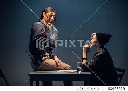 A young asian female investigator sits on a table and shows a package of weapons to a blonde suspect A young asian female investigator sits on a table and shows a package of weapons to a blonde suspect 80477319