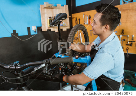Handsome bicycle mechanic in an apron wearing gloves when installing a saddle 80480075