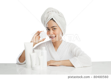 Smiling beautiful girl model with towel on head holding cream pot bottle with various skin care products on table Smiling beautiful girl model with towel on head holding cream pot bottle with various skin care products on table 80480560