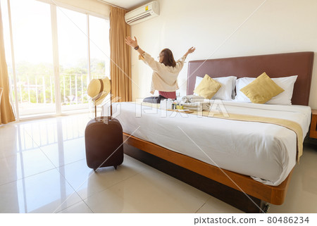 Portrait of tourist woman sitting on the bed, looking to beautiful view outside the window with her luggage in hotel bedroom after check-in. Conceptual of travel and vacations. Portrait of tourist woman sitting on the bed, looking to beautiful view outside the window with her luggage in hotel bedroom after check-in. Conceptual of travel and vacations. 80486234