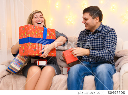 portrait of adult romantic couple, sitting with gifts on a couch at home, a woman takes away all the gifts from a man, the photo is like a joke, holiday lights on a wall 80492308