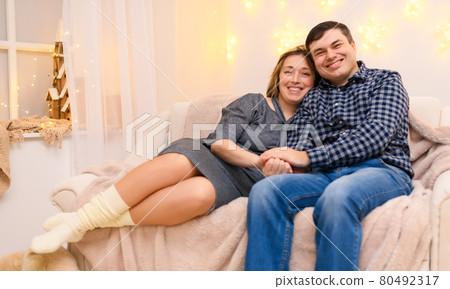 portrait of adult romantic couple sitting on a couch at home, holiday lights on a wall portrait of adult romantic couple sitting on a couch at home, holiday lights on a wall 80492317