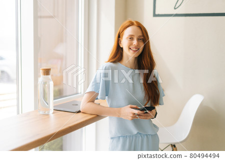 Medium shot of smiling charming young woman holding mobile phone, typing online messages standing by window in cozy light cafe. 80494494
