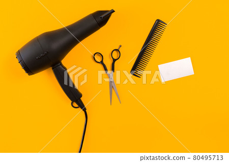 Black hair dryer, comb and scissors on yellow paper background. Top view 80495713