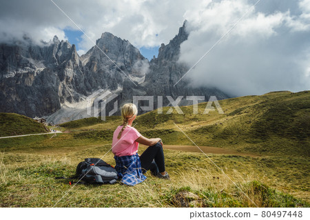 Dolomites. Woman sitting on the grass enjoying Baita Segantini mountain with Cimon della Pala peak, refuge and lake in background. Rolle pass, Trentino province, Italy, Europe 80497448