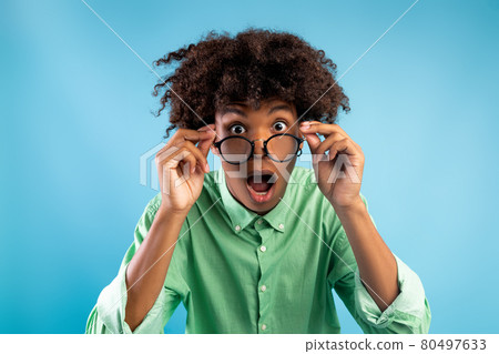 Emotional african american teen guy in glasses opening mouth in surprise, looking at camera over blue background Emotional african american teen guy in glasses opening mouth in surprise, looking at camera over blue background 80497633