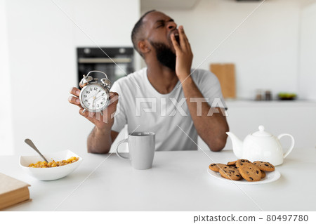 Black man suffering from insomnia yawning, holding showing alarm clock 80497780