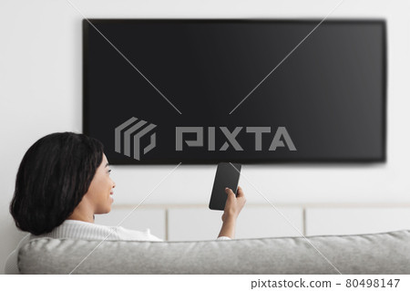 Smart home entertainment controlled by smartphone app. Happy woman with cellphone connected to TV, empty space Smart home entertainment controlled by smartphone app. Happy woman with cellphone connected to TV, empty space 80498147