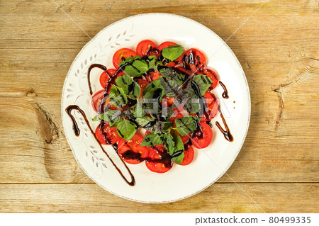 Cut red cherry tomatoes and basil leaves on white plate with balsamic vinegar sauce, wood background 80499335