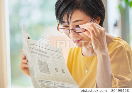 A middle-aged woman wearing reading glasses and reading an English newspaper A middle-aged woman wearing reading glasses and reading an English newspaper 80499468