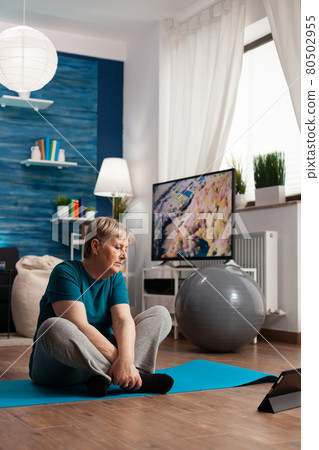 Retired senior woman in sportswear sitting on yoga mat watching fitness lesson on laptop Retired senior woman in sportswear sitting on yoga mat watching fitness lesson on laptop 80502955