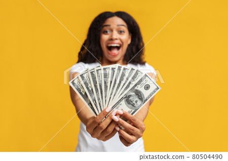 Overjoyed black woman holding money cash in hands, standing over yellow studio background, selective focus 80504490