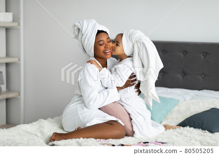 Full length portrait of little girl kissing her black mother on makeup day, wearing bath robes and towels in bedroom 80505205