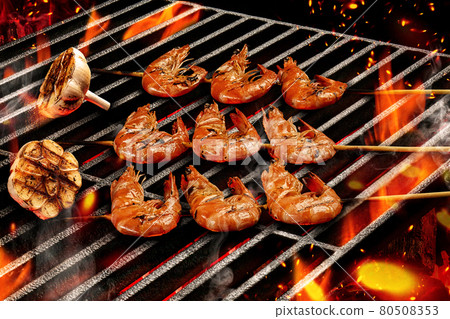 Barbecue bbq grilled delicious roasted shrimps on wooden skewers with halves of garlic. Flaming fire and ember charcoal. Seafood, shellfish. Close up Barbecue bbq grilled delicious roasted shrimps on wooden skewers with halves of garlic. Flaming fire and ember charcoal. Seafood, shellfish. Close up 80508353