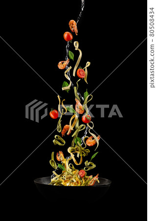 Wok pan, falling pasta with tomatoes, shrimps and green basil against black studio background. Poured by a water stream. Cooking concept. Close up Wok pan, falling pasta with tomatoes, shrimps and green basil against black studio background. Poured by a water stream. Cooking concept. Close up 80508434