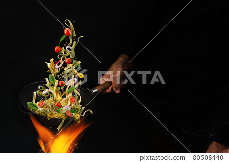 Unrecognizable man holding wok pan above fire and cooking pasta with cherry tomatoes, onion and basil against black background. Close up 80508440