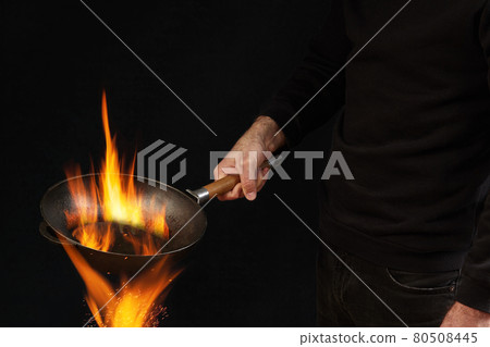 Young guy dressed in jumper and jeans. He is holding burning wok pan above fire, against black studio background. Cooking concept. Close up Young guy dressed in jumper and jeans. He is holding burning wok pan above fire, against black studio background. Cooking concept. Close up 80508445