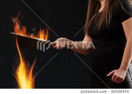Brunette girl with tattooed hands, dressed in leggings and t-shirt. Holding burning wok pan above fire against black background. Cooking. Side view Brunette girl with tattooed hands, dressed in leggings and t-shirt. Holding burning wok pan above fire against black background. Cooking. Side view 80508454