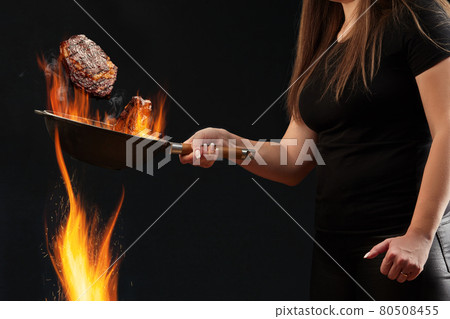Woman with tattooed hands, dressed in leggings and t-shirt. Holding burning wok pan above fire and frying two beef steaks, black background. Side view Woman with tattooed hands, dressed in leggings and t-shirt. Holding burning wok pan above fire and frying two beef steaks, black background. Side view 80508455