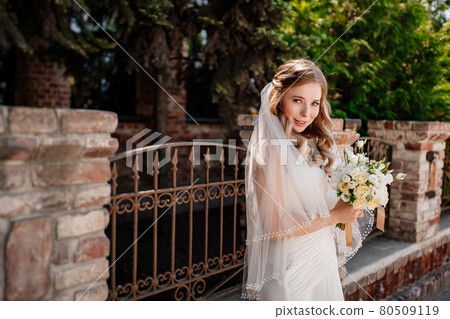 gentle and beautiful bride in a veil with a bouquet of flowers at the fence. 80509119