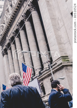 Businessman talking on the phone on Wall street in New York with American flags and New York Stock Exchange in background. 80510524