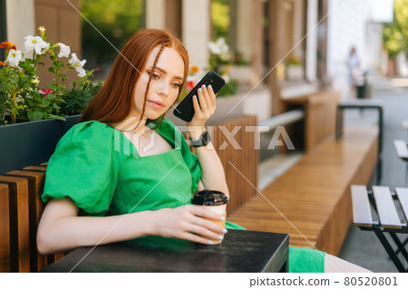 Pretty young woman wearing casual green dress listening voice message from smartphone sitting at table in outdoor cafe terrace in sunny summer day. Pretty young woman wearing casual green dress listening voice message from smartphone sitting at table in outdoor cafe terrace in sunny summer day. 80520801