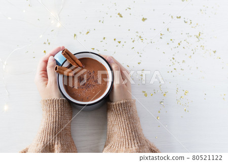 Hot chocolate with cinnamon spice in enamel mug in female hands on white wooden background top view. 80521122